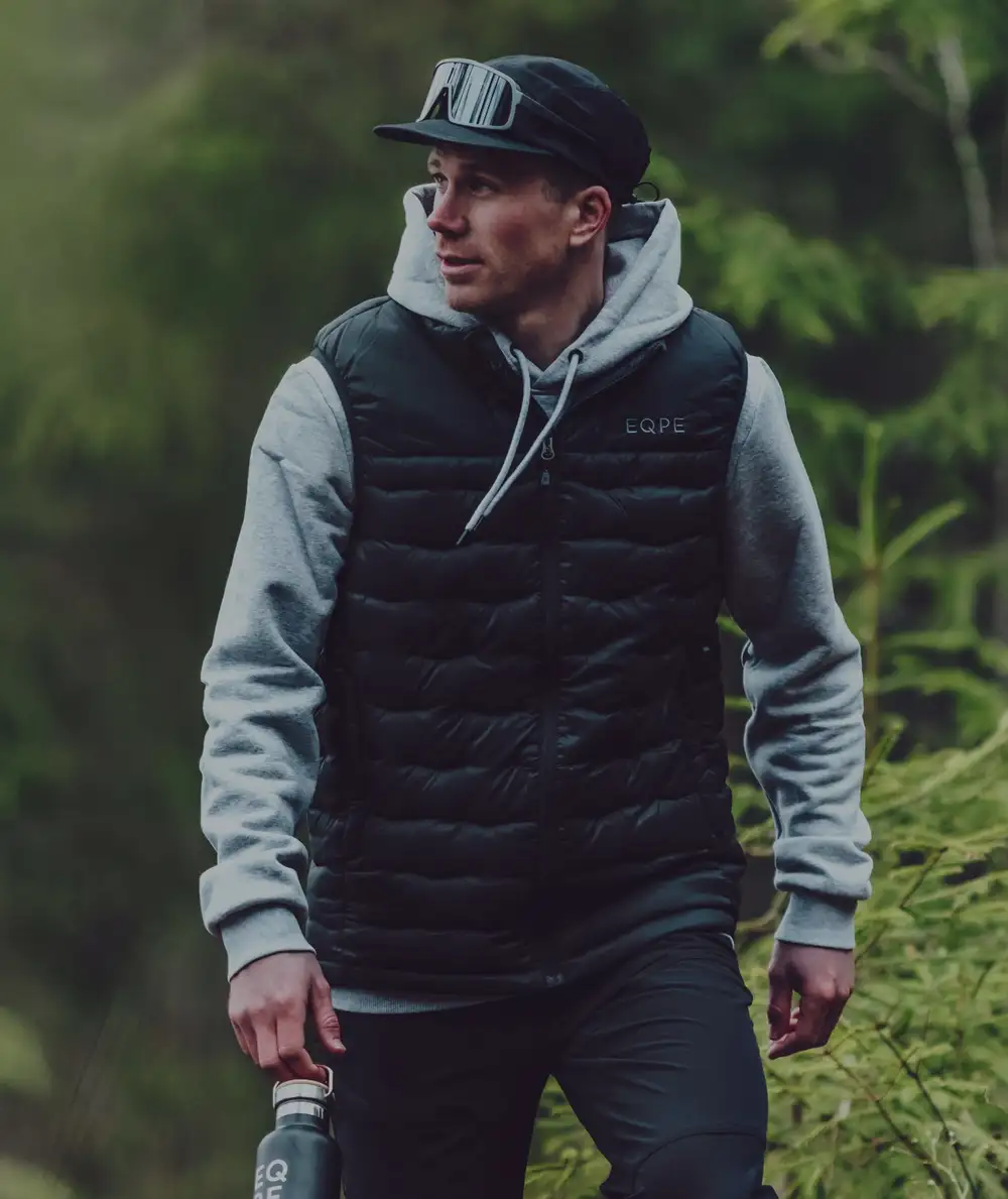 Man in a black cap, puffer vest, and gray hoodie walks in a forest.
