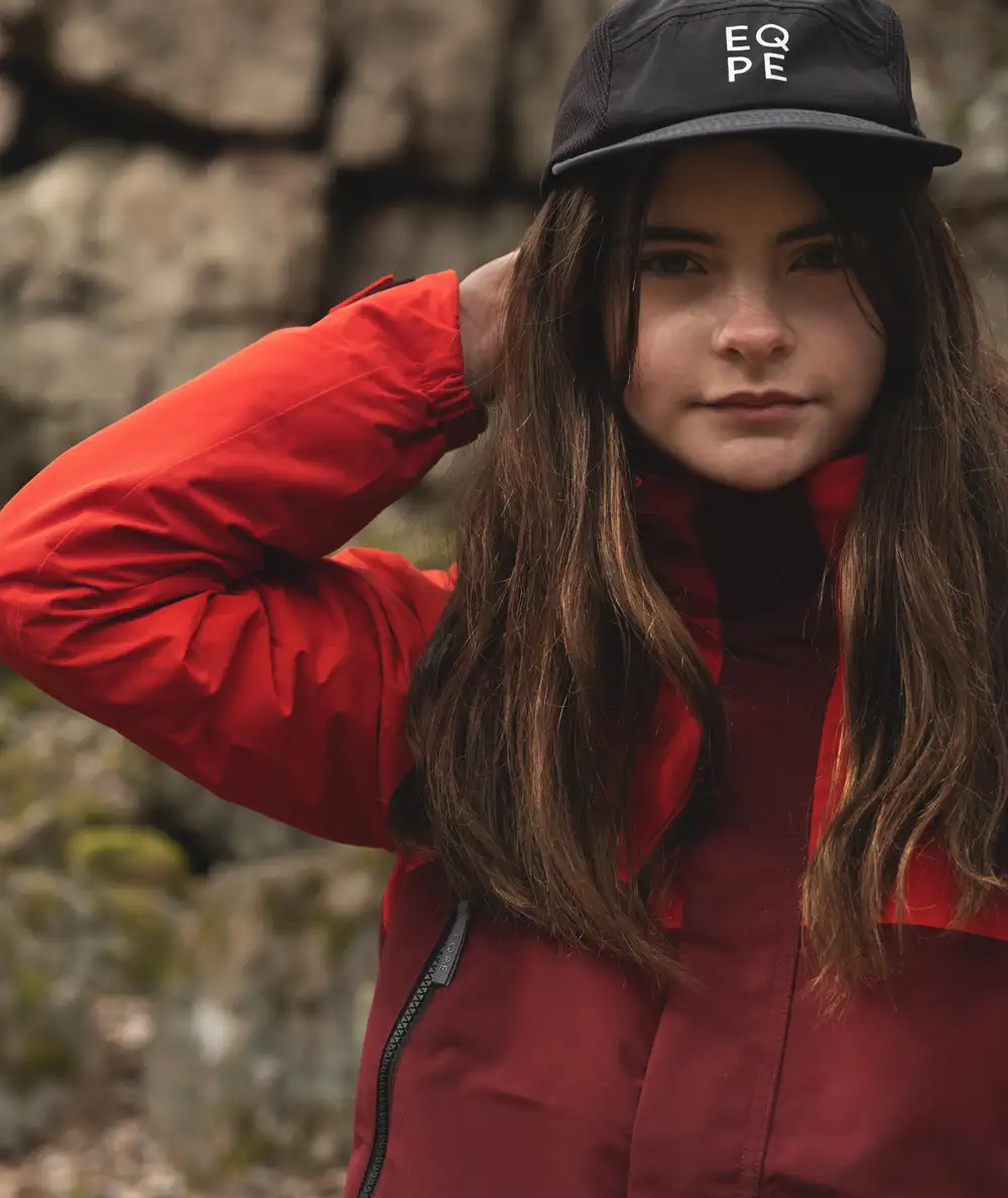 A young girl in a red jacket and black cap stands outdoors against a rocky background.