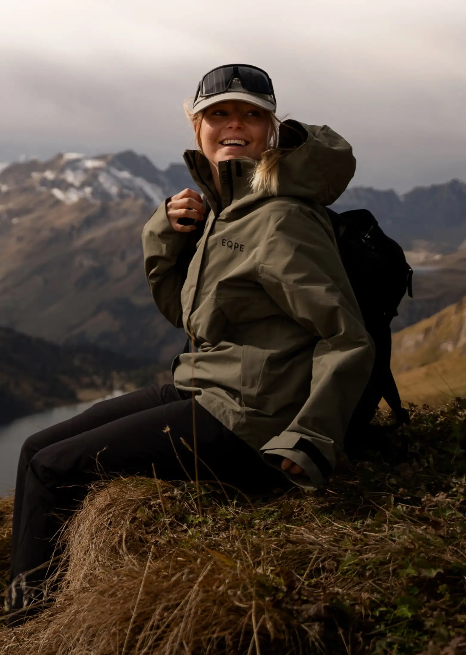 A smiling woman wearing an olive-green outdoor jacket, black pants, and a cap with goggles sits on a grassy mountain ridge with a black backpack. Snow-capped peaks and a lake are visible in the background under a cloudy sky.