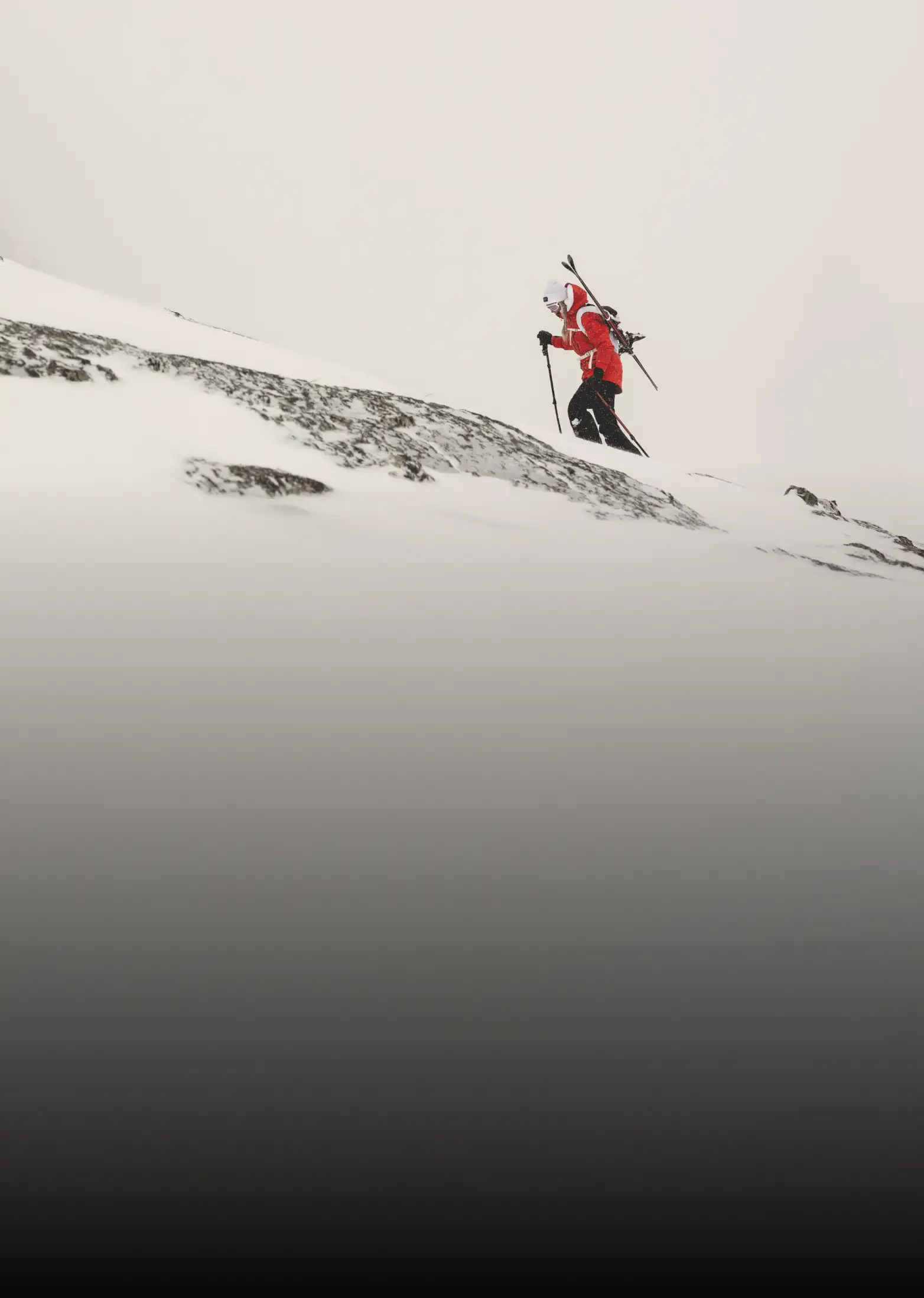 A person in a red jacket and black pants hikes up a snow-covered mountain slope with skis strapped to their back and poles in hand, surrounded by fog or heavy snowfal.