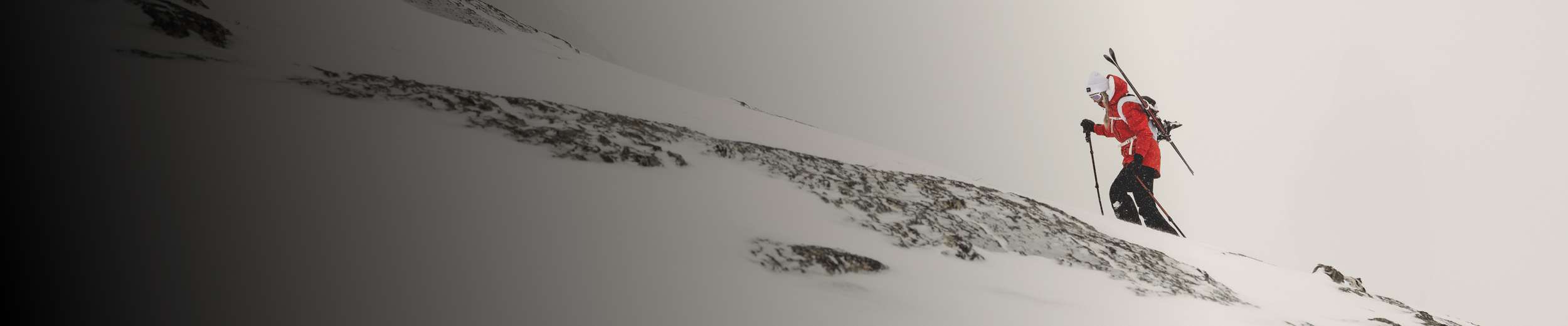 A person in a red jacket and black pants hikes up a snow-covered mountain slope with skis strapped to their back and poles in hand, surrounded by fog or heavy snowfal.