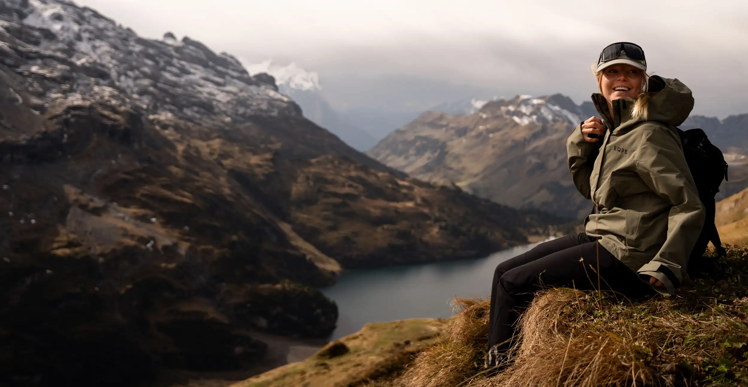 A smiling woman wearing an olive-green outdoor jacket, black pants, and a cap with goggles sits on a grassy mountain ridge with a black backpack. Snow-capped peaks and a lake are visible in the background under a cloudy sky