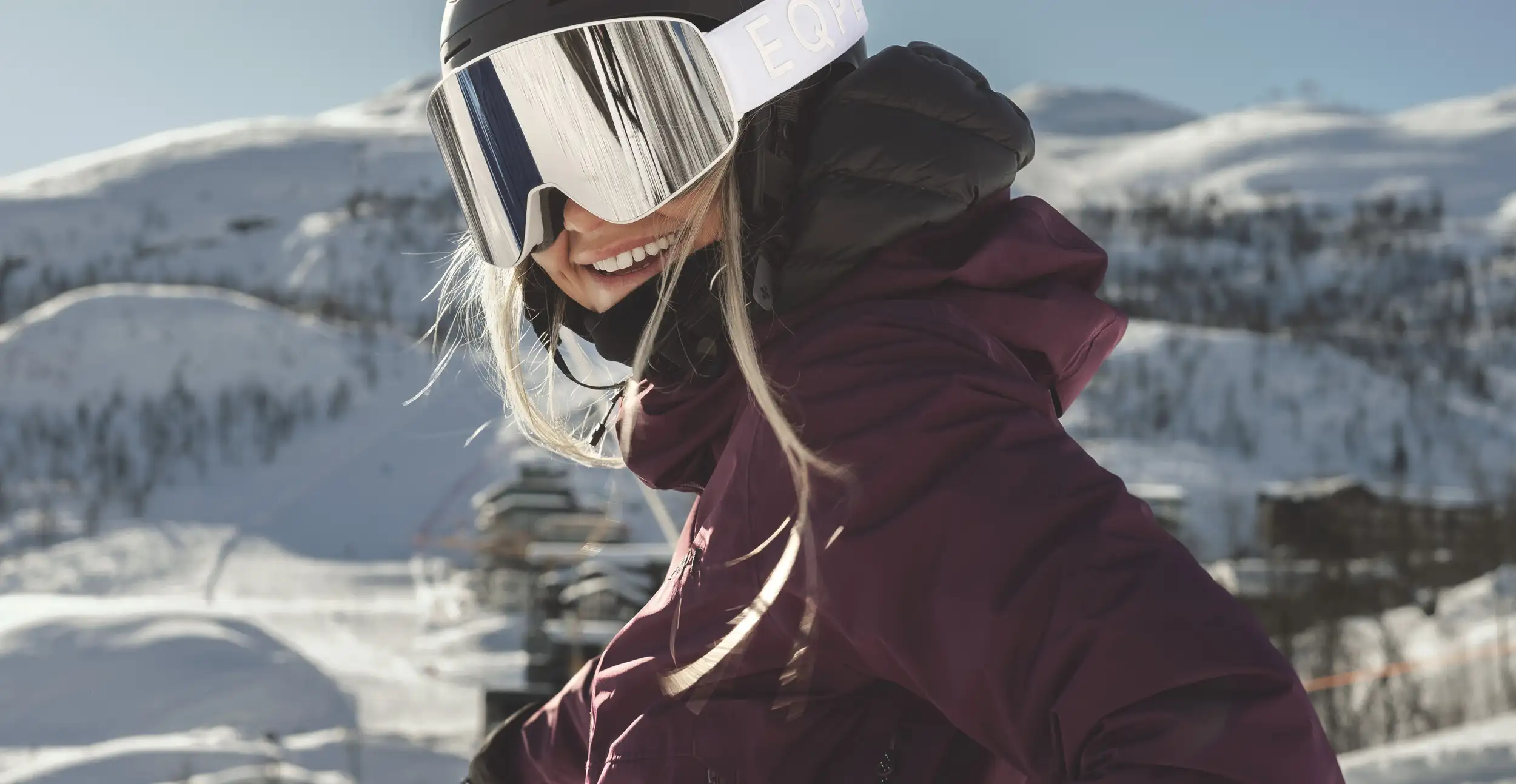 A smiling woman wearing a black ski helmet, mirrored ski goggles with "EQPE" branding, and a dark purple ski jacket, standing in a snowy mountain setting.