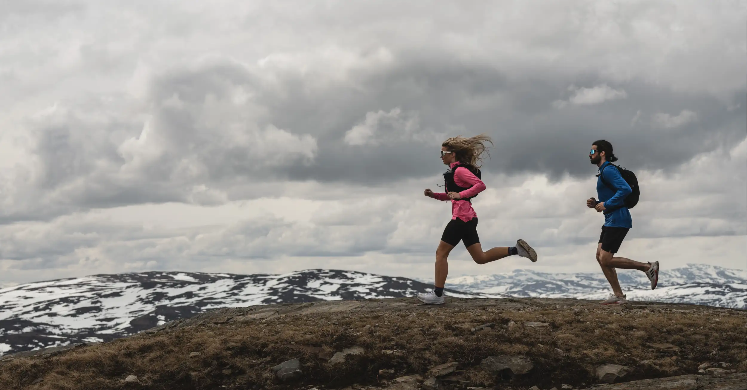 Two people running on a rocky mountain trail, wearing athletic gear under a cloudy sky.